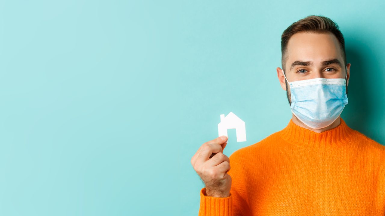 Real estate and coronavirus pandemic concept. Close-up of adult man in medical mask holding small paper house maket and smiling, searching for apartment, light blue background.