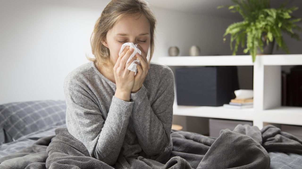 Sick young Caucasian woman covered with grey blanket sitting on bed with closed eyes, blowing nose with napkin. Illness, pain concept